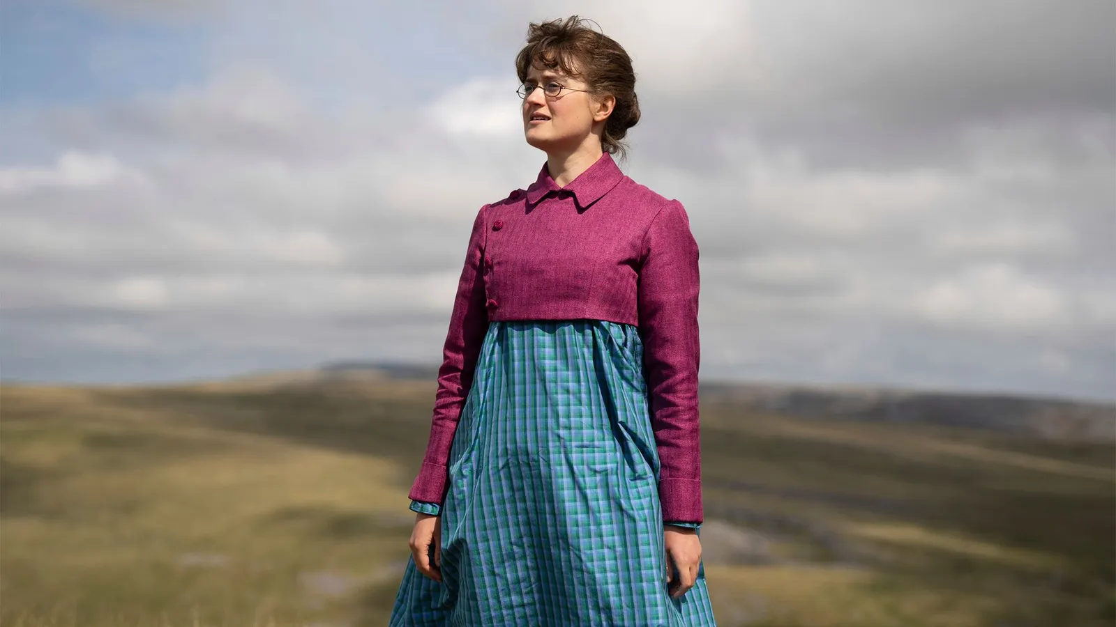 A young woman in front of dramatic rolling hills with a cloudy sky above her stares into the distance alone.